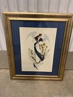 Framed needlework art of an angel wearing a blue and white flowing dress with a golden ribbon, white wings, mounted in a golden textured frame with dark blue matte backing.