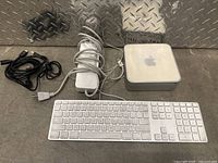Apple Mac Mini, keyboard, and power adapter with cables on metal surface.