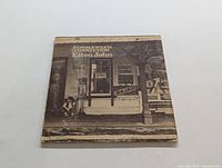 Front cover of Elvis John's 'Tumbleweed Connection' LP showing a sepia-toned vintage street scene with signage and a person sitting on the front porch
