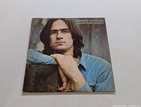 Front cover of James Taylor 'Sweet Baby James' vinyl LP showing a portrait of the artist leaning on a wooden surface wearing a blue shirt.