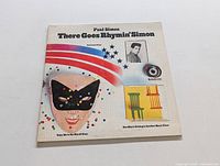 Front cover of vinyl LP 'There Goes Rhymin' Simon' by Paul Simon showing album title and artistic illustrations including a masked face and rainbow stripes.