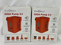 Two boxed MicroNovelty GIGA Pump 3.0 portable air pumps in original sealed packaging. The boxes show the product image in bright orange with multifunction icons for air pump, vacuum pump, and lantern modes, and nozzle attachments.