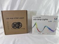 Two boxes of LED RGB strip light kits; one Luxense S1 model in white packaging, another generic kit in brown box with spool image.
