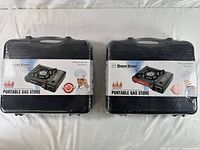 Two black plastic carrying cases containing Happy Home portable gas stoves, shown side-by-side, sealed in plastic wrap.