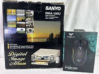 Front view of Sanyo DMA-100U Digital Image Album converter box next to AULA FG06 RGB gaming mouse box showing packaging and branding.