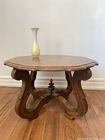 Front view of octagonal antique Victorian style solid wood side table with curved legs and central finial base, placed on hardwood floor with cream wall background. Cream vase on table top for scale.