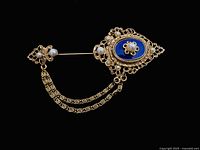 Front view of gold-tone stick pin brooch with oval blue enamel center, filigree detailing and faux pearl accents, attached to a smaller pin by two linked chains.