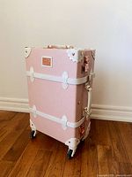 Photo showing front and one side of pink embossed carry-on luggage with white trim and rose gold hardware on wooden floor against white wall.