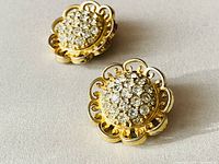 Pair of gold tone clip-on earrings with a circular rhinestone cluster center and scalloped openwork edge, on light fabric background.