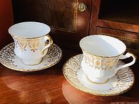 Pair of white Paragon porcelain teacups and matching saucers with intricate gold gilt decoration on wooden surface.