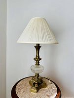 Full view of the vintage brass and crystal base lamp with pleated lampshade against a neutral wall and on a round marble table.