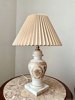 Full view of vintage French white glass table lamp with gold detailing and beige pleated lampshade standing on a pink marble topped table.
