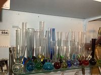 Front view of assorted art glass bud vases and bar glasses on glass shelf, showing colorful rounded bases with bubbles, varying heights up to approx. 10 inches.