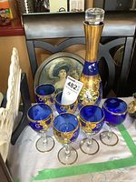 Photo showing the cobalt blue decanter with stopper surrounded by six decorated glasses, set on a table with a white cloth and green tape edge.