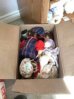 Top view of open box filled with assorted dolls inside, showing doll dressed in plaid and floral outfits