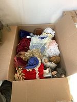 Full box view showing multiple porcelain dolls dressed in period style costumes with lace and ribbons, some laying down