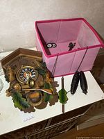 Vintage carved wood cuckoo clock shown with detached pendulum and two pinecone-shaped weights, placed near a pink fabric basket.