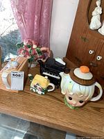Photo showing all five unique ceramic teapots arranged on a wooden surface near a pink curtain, including strawberry basket, black piano, cottage house, white child head, and smaller biscuit lid teapots.