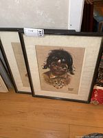 Two framed prints with black frames and cream mats showing portraits of a young child with necklaces and braids, signed by artist.