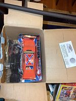 Top-down photo of the 1969 General Lee die cast model kit parts packed inside a cardboard inner box, showing a bright orange model car with roof markings and separate chassis part.