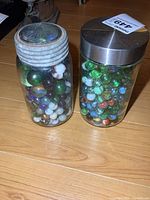 Two glass jars filled with assorted marbles with different lid types on wooden floor
