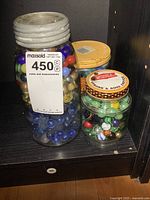 Three glass jars on a shelf containing multiple colorful marbles with one labeled maxsold 450 tag attached.