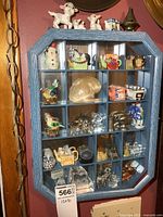 Front view of small blue wooden display case showing various miniatures in 24 compartments and three ceramic dog/cat figurines alongside a ceramic house and metal lantern on top.