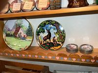 Shows two 12-inch round paintings on a shelf: one with a cottage and ducks, another convex with deer, plus two stacks of porcelain coasters.