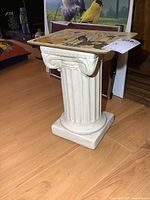White ceramic pedestal shaped like a classical fluted column on wooden floor.