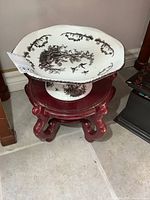 View of large porcelain pedestal dish on red wooden stand on tiled floor.