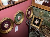 Four oval framed porcelain plaques hung on a red wall, showing detailed hand-painted romantic scenes with gold ornate frames and velvet mats in burgundy, green, and black.