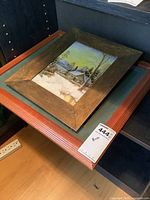 Larger wooden framed print showing a snow-covered cabin with trees and a yellowish sky.