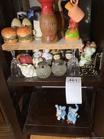 Two shelves featuring multiple vintage salt and pepper shakers including hamburger pairs, wooden lady figure, colorful rooster, glass and ceramic shakers, and bunny set below.