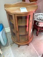 Front and angled side view of small wooden curio cabinet showing curved glass panels and door with lock.