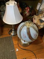 Photo showing both table lamps side by side on a wooden floor. Left lamp has white fabric shade and metal base. Right lamp has round glass shade with floral decorations and gold accents, and gold-tone base.