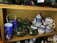 Four teapots with matching cream, sugar pots and cups arranged on a shelf showing dark green set, blue Japanese-style cups, and purple-blue and white ceramic pieces