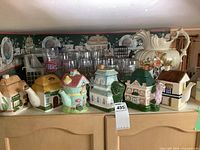 Full view of the six ceramic teapots lined up on top of a wooden cabinet with glasses behind. Each teapot is in the shape of a different house with various colors and details.