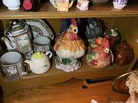Shelf view showing multiple teapots and mugs including rooster and hen designs, a Fitz and Floyd hen teapot, smaller teapots, and a decorative plate