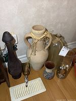 Photo showing large beige ceramic vase, tall olive green glass bottle, dark metal decanter, small floral vase, clear glass vase with decorative balls, and brass wall candle holder.