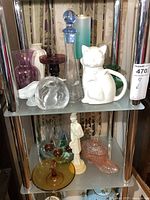 Two shelves containing small glass vases in various colors, figures including a white cat ceramic pitcher, a clear glass decanter with stopper, and other small decorative glass pieces.
