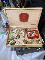 Open wooden cigar box showing assortment of small Wade ceramic figurines and metal thimbles in small red box.