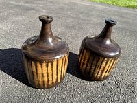 Photo of two medium vintage brown wood bottles with vertical striped pattern, cylindrical body and narrow necks, outdoors on asphalt surface showing overall condition and coloration.