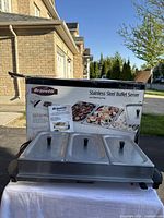 Front view of Bravetti stainless steel buffet server with three lidded trays and original box behind it.