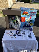 Buzz Lightyear telephone in original box with handset, cord, multilingual instructions and warranty documents on white cloth with outdoor background.