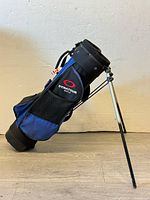 Side view of a blue and black Dynatour golf stand bag with attached dual metal legs for standing support, placed against a white wall and wooden floor.