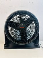 Front view of black Honeywell table fan showing the protective grill, fan blades, and brand logo on the central hub. Fan is powered by a cord seen at the back.