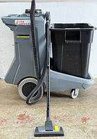 Side view of gray plastic cleaning cart with large wheels, vacuum hose attached, and black trash bin on back shelf. Vacuum head with yellow detail in front.