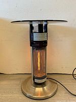 Front view of the infrared patio heater showing metal mesh covering and glowing heating element with round base, glass table top above it.