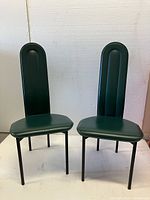 Front view of two green upholstered antique chairs with tall backrests and black metal legs.