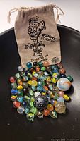 Burlap bag laid out with many assorted marbles in front showing various colors, patterns, and sizes, including some larger marbles with space-like swirled designs.